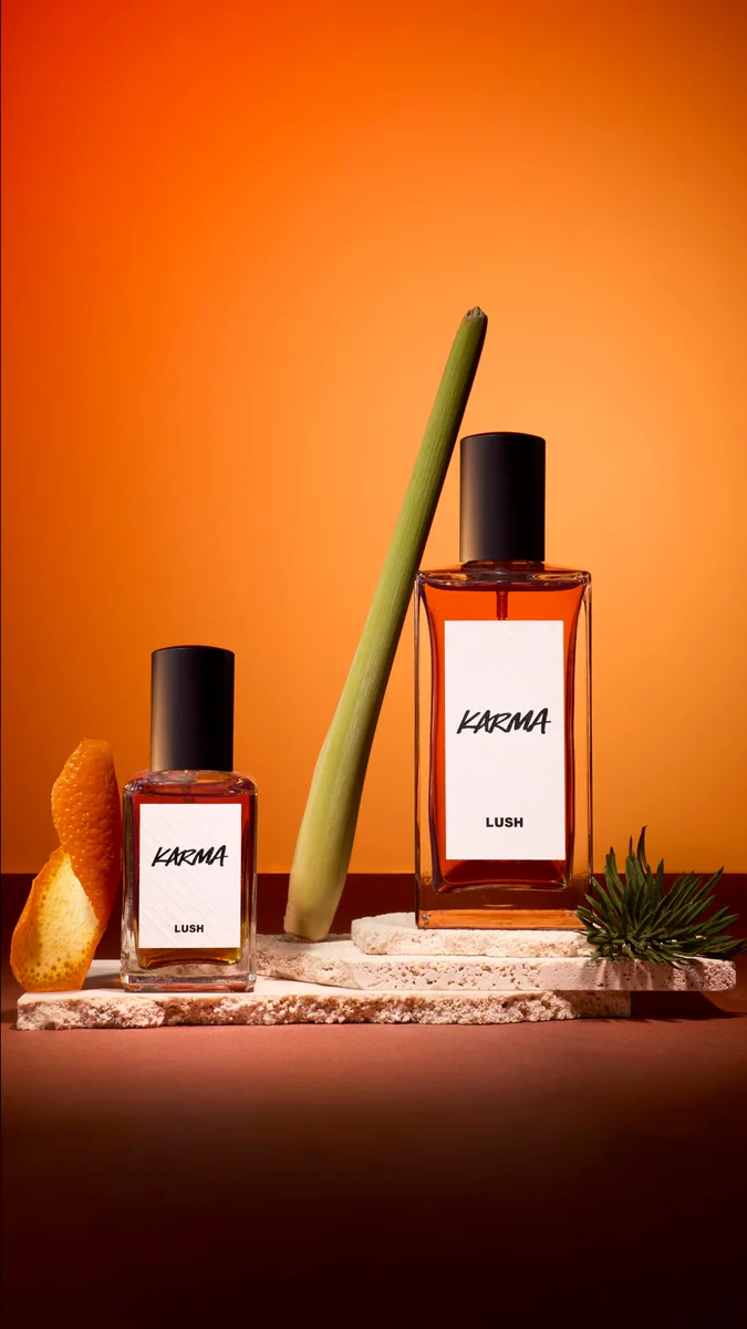 Two bottles of Lush Karma perfume on stone slabs against an orange background, styled with orange peel, lemongrass and pine
